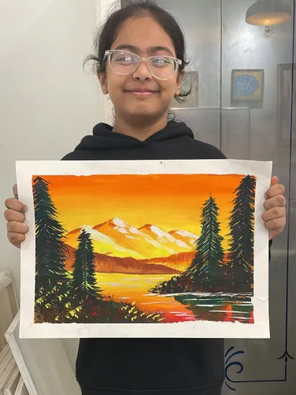 A young student holds up her poster color landscape painting of a mountain sunset. She did a wonderful job blending the warm colors of the sky and reflecting them in the water.