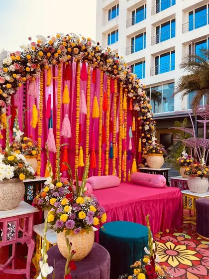 A beautiful seating area for a Mehendi ceremony, featuring a pink diwan decorated with colorful tassels and surrounded by vibrant floral arrangements.