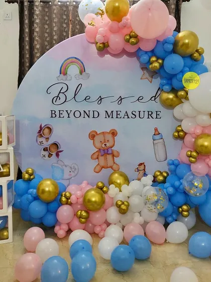 An intimate baby shower setup perfect for a home celebration. This "Blessed Beyond Measure" backdrop features a cute teddy bear and rainbow design, framed by a classic pink and blue balloon garland.