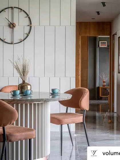The dining area is defined by a fluted pedestal table with a marble top, paired with contemporary upholstered chairs. The vertically paneled wall and oversized clock add a sense of height and simple elegance to this compact dining nook.