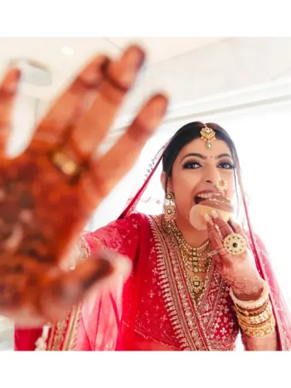 A fun and candid shot of the bride enjoying a snack while getting ready, playfully showing her mehendi-adorned hand to the camera. It's a real moment amidst the wedding glamour.