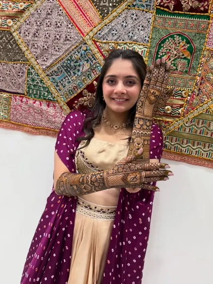 A full-view shot of the bride against a beautiful, colorful tapestry. Her intricate mehndi, filled with personal stories, is the perfect accessory for her traditional outfit.