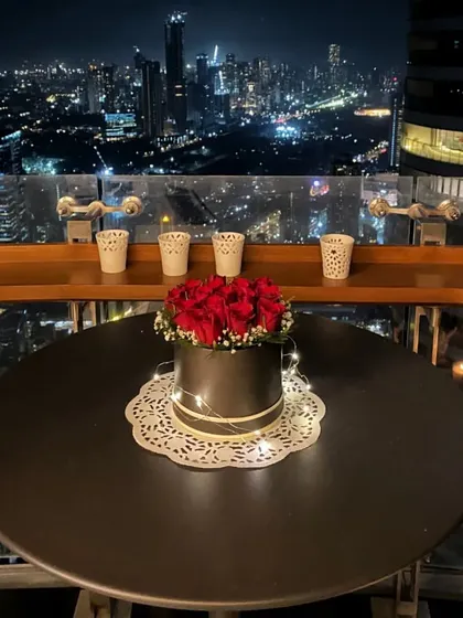 A nighttime view of the romantic table setup. The box of red roses, illuminated by fairy lights, becomes the glowing centerpiece against the sparkling city skyline.