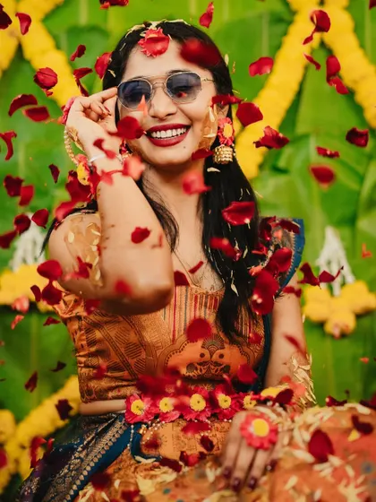The bride smiles, her face adorned with floral jewelry and a shower of red petals during her Haldi ceremony.