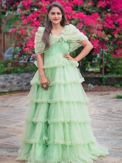 A model wearing a pastel green tiered ruffle gown. This dress has a beautiful, voluminous silhouette and comes with a matching ruffled capelet.