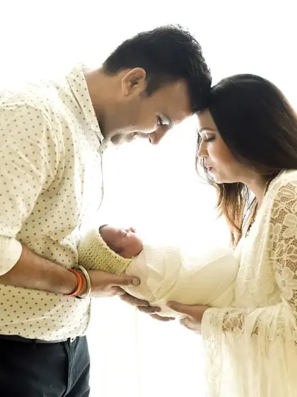 A tender moment captured against a bright, clean background. The parents hold their newborn together, their heads touching in a gesture of unity.