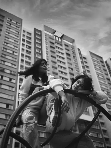 A dramatic, low-angle black and white shot of two women on a playground structure against a backdrop of towering apartment buildings. The urban setting and composition create a powerful, modern image.