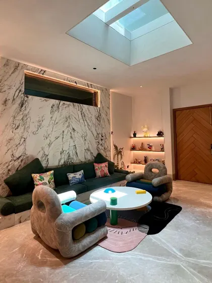 This wide view shows how the playful furniture interacts with the architectural elements of the room. A large skylight illuminates the space, highlighting the dramatic marble feature wall and the custom built-in sofa, creating a bright and dynamic living area.