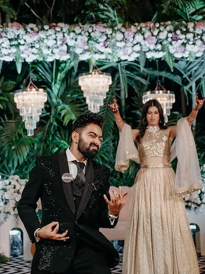 The couple performs on their modern sangeet stage, framed by a floral arch and hanging crystal chandeliers.