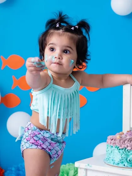 Offering a little taste of the delicious cake. The bright blue background and her mermaid-themed outfit make the colors pop, creating a vibrant and joyful image.