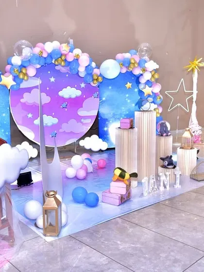 A dreamy 'Star and Moon' themed party setup. The decor features a night-sky backdrop with cloud and star cutouts, a crescent moon prop, and a beautiful balloon arrangement in shades of blue, pink, and gold.