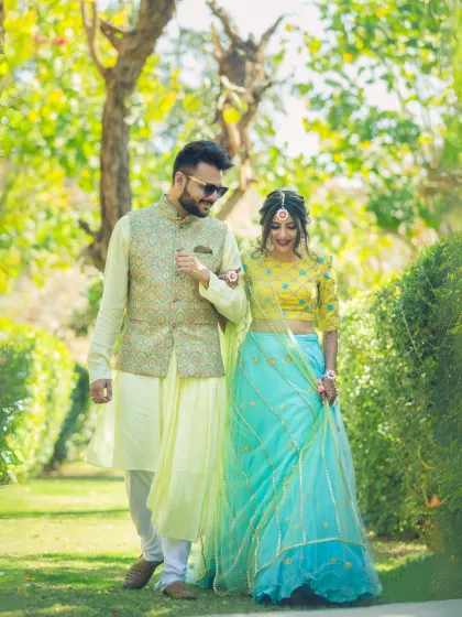 A happy couple takes a stroll through a lush green garden. Their coordinated, colorful outfits for a Mehendi or Haldi ceremony make this a vibrant and cheerful pre-wedding portrait.