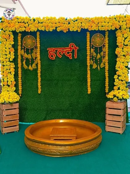 A simple yet beautiful Haldi decoration with a green grass backdrop, a frame of marigold flowers, and a neon Hindi "Haldi" sign. The wooden crates and dreamcatchers add a rustic, bohemian touch to the setup.