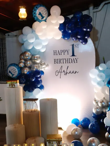 I can create beautiful and simple decorations for parties at home. This is a clean and modern setup for a first birthday, using a balloon arch in blue, white, and silver against a personalized backdrop.