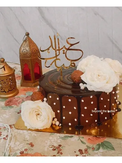 An elegant chocolate drip cake for Eid, decorated with white roses and a beautiful Arabic "Eid Mubarak" topper.