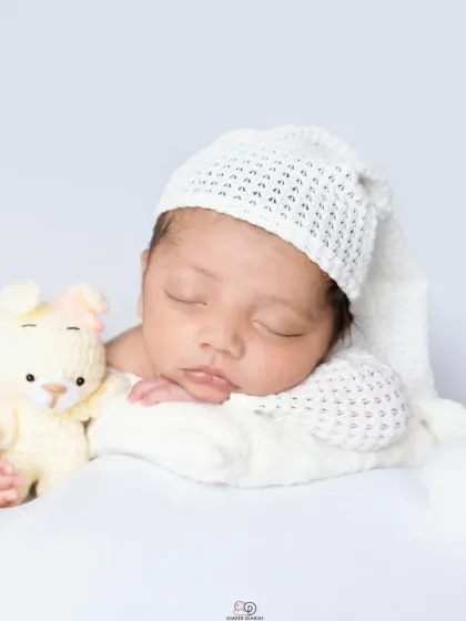 A pure and simple newborn portrait against a clean white background. The baby is dressed in a white knitted outfit and sleepy hat, showcasing their angelic features.