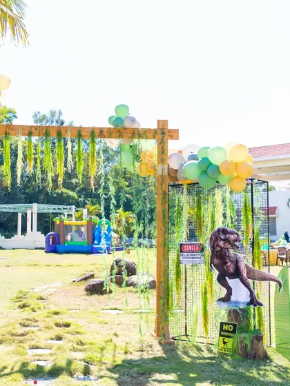 The entrance to the dinosaur park, featuring a caged T-Rex and a bouncy castle in the background, setting the scene for a day of adventure.