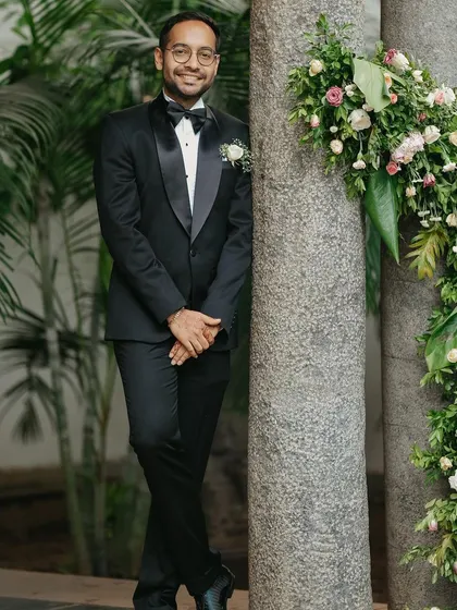 A dapper portrait of the groom in his reception tuxedo, standing next to a pillar adorned with flowers.