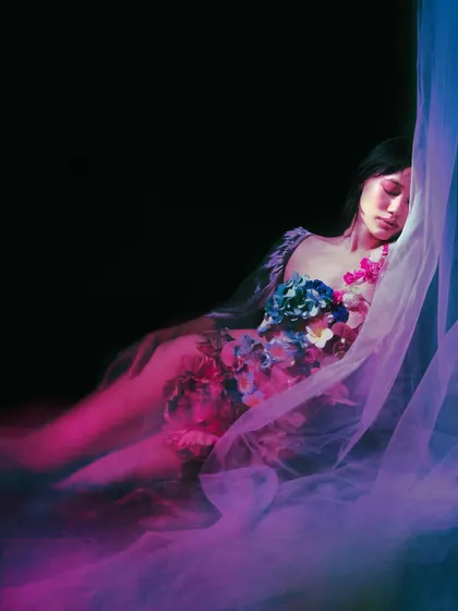 The model lies in a haze of purple and pink smoke or fabric, eyes closed, reminiscent of Ophelia floating in water. The dramatic colors and soft focus lend a surreal, otherworldly feel to the scene.