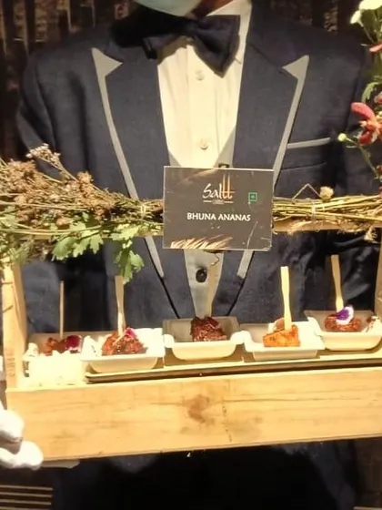 Our server presents a tray of Bhuna Ananas, a smoky and sweet grilled pineapple appetizer. The rustic wooden tray adorned with fresh flowers and herbs demonstrates our unique and thoughtful presentation style.
