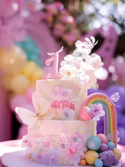 A beautiful two-tiered cake for Ayra's first birthday, decorated with delicate butterflies, a rainbow, and pastel-colored spheres.