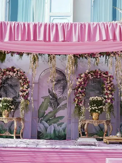 The elegant stage setup, featuring a backdrop of hand-painted panels with tropical leaf motifs. Arches of pink and white flowers frame the seating area, creating a perfect photo opportunity.