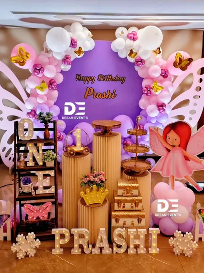 A grand butterfly-themed first birthday for Prashi. The setup includes large butterfly wing backdrops, a purple balloon arch, and a full display of themed props and marquee letters.
