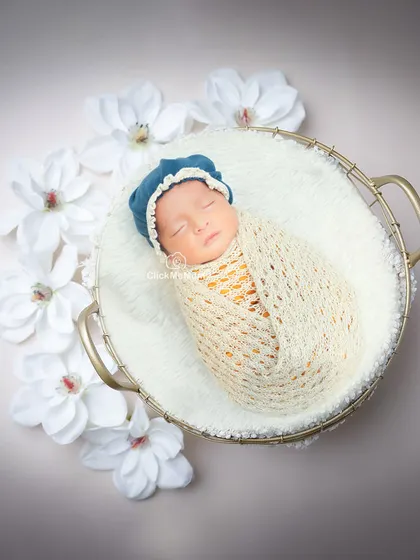 This baby boy, in a cream-colored swaddle and blue bonnet, looks like a precious pearl surrounded by white flowers in a golden basket.