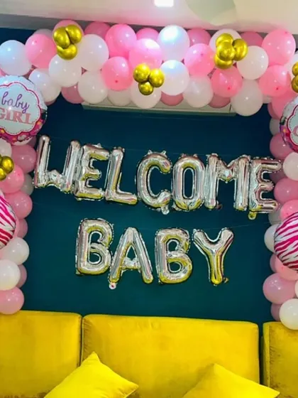 A "Welcome Baby" girl decoration set up over a sofa. The balloon arch in pink, white, and gold perfectly frames the silver foil letter balloons.