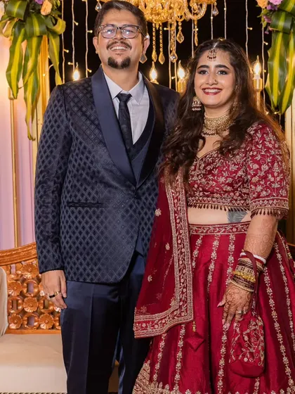 The couple posing for a photo. Her hair is styled in soft waves, and her makeup is a perfect match for her elegant red lehenga.