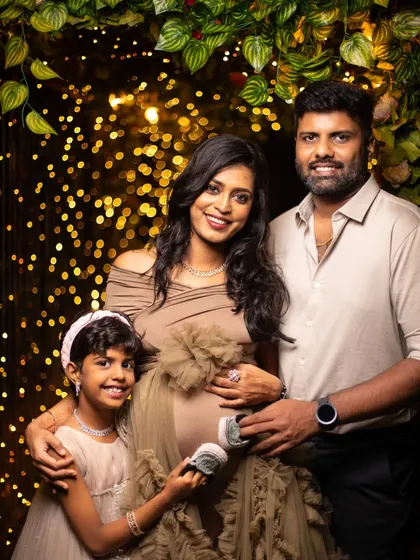 A modern studio maternity portrait with a touch of glamour. The family is framed by a backdrop of warm bokeh lights, with their daughter holding tiny shoes in anticipation of her new sibling. It's a stylish and heartfelt family photo.