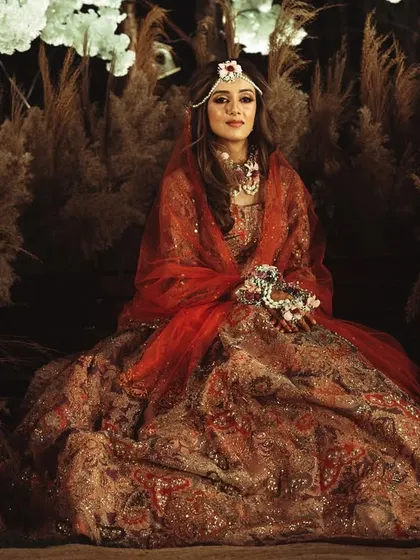 A regal portrait of the bride seated amidst pampas grass decor. Her elaborate red lehenga and direct gaze create a powerful and stunning image.