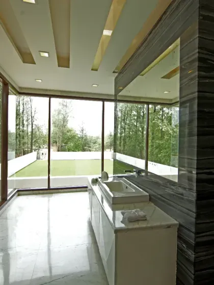 The master bathroom at Sachdeva Farmhouse is a light-filled space with floor-to-ceiling glass offering views of the pool and gym terrace. A sleek stone feature wall screens the sunken tub, creating a refined, spa-like feel.