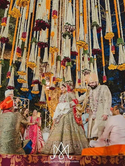 A sacred wedding ritual takes place on a stage elevated with traditional fabrics and surrounded by a canopy of hanging floral tassels, creating a divine atmosphere.
