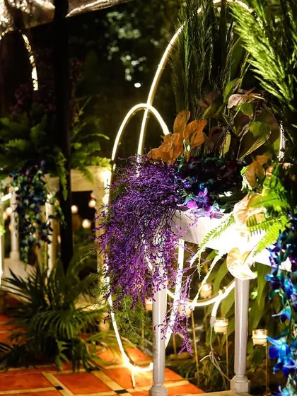The entrance pathway to the Diwali party, lined with lush greenery and vibrant purple and blue floral arrangements. The modern circular light elements add a contemporary touch.