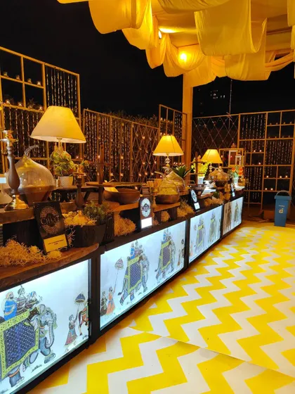 A vibrant food counter with an illuminated front panel depicting a royal Indian procession. The yellow and white chevron flooring and golden decor create a festive and modern take on a traditional Indian mela theme.