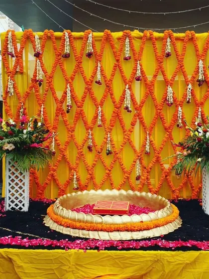 A simple and traditional setup for a Haldi ceremony. The backdrop is made of a lattice of orange marigold garlands, creating a classic and vibrant look.