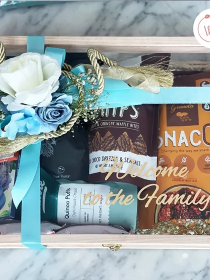 A "Welcome to the Family" hamper in a clear-lid wooden box. This gift includes a mix of healthy and indulgent snacks, perfect for making a new family member feel right at home.