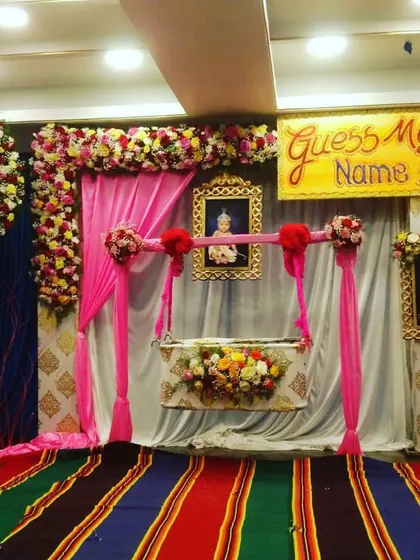 A beautiful naming ceremony setup featuring a floral-decorated swing cradle as the centerpiece. The design includes a large floral ring with the baby's photo and a "Guess My Name" sign for a playful touch.