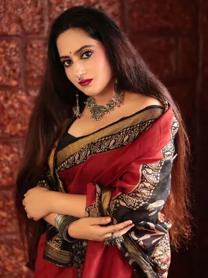 A striking portrait of a model in a red and black saree. Her direct gaze and the way she holds her arm create a strong and confident pose, highlighting her bold jewelry.
