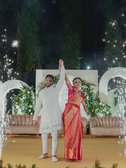 The couple celebrates their union on a modern, minimalist stage with clean white arches, lush green foliage, and celebratory cold pyro fireworks.