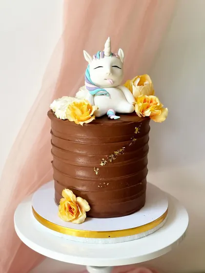 A fun and cheeky "fat unicorn" cake, featuring a sleepy unicorn resting on top of a rich chocolate buttercream cake. Yellow sugar flowers and gold leaf add a touch of whimsy.