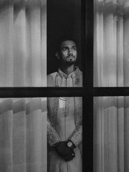 A dramatic black and white portrait of a groom seen through a window. The play of light and shadow creates a moody and cinematic feel.