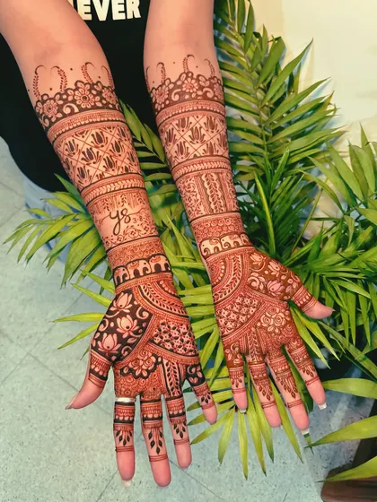 A stunning full-arm design set against a natural background, showing how the rich henna color complements any setting. This piece includes personalized initials and a mix of floral and geometric bands.
