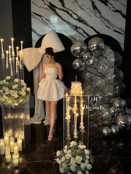 A glamorous birthday girl poses within her stunning party setup. The decor features a dramatic white bow, elegant candelabras, and a backdrop of silver disco balls, creating a high-fashion, celebratory atmosphere.