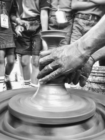 A black and white photo showing an expert potter demonstrating the craft to a group of curious children, passing on a timeless skill.