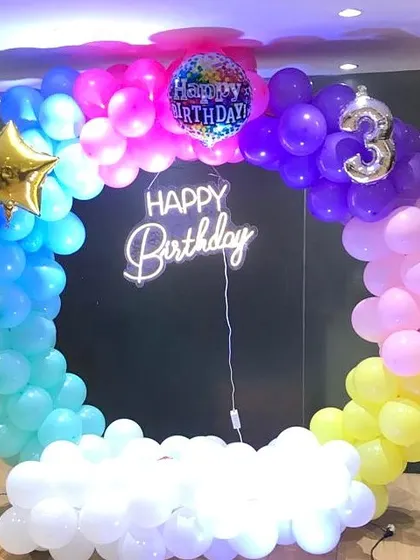 A colorful 3rd birthday photo booth with a rainbow-themed circular balloon arch and a cloud base made of white balloons.