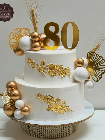 This two-tier cake was designed for a grand 80th birthday celebration. I used a clean white whipped cream frosting as the canvas for elegant gold leaf, decorative spheres, and a bold topper, creating a sophisticated and memorable design.