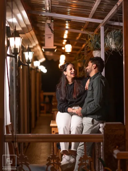 A relaxed evening moment on a beautifully lit verandah. The warm string lights create a cozy and inviting atmosphere for Ishu and Arihant's shared laughter, showing that romantic shots can also be casual and comfortable.