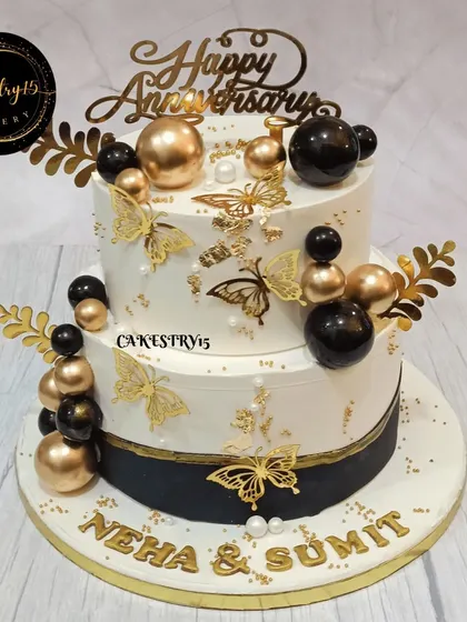A luxurious two-tier choco-vanilla anniversary cake. The design features a black and gold theme with golden butterflies, decorative balls, and a custom nameplate.
